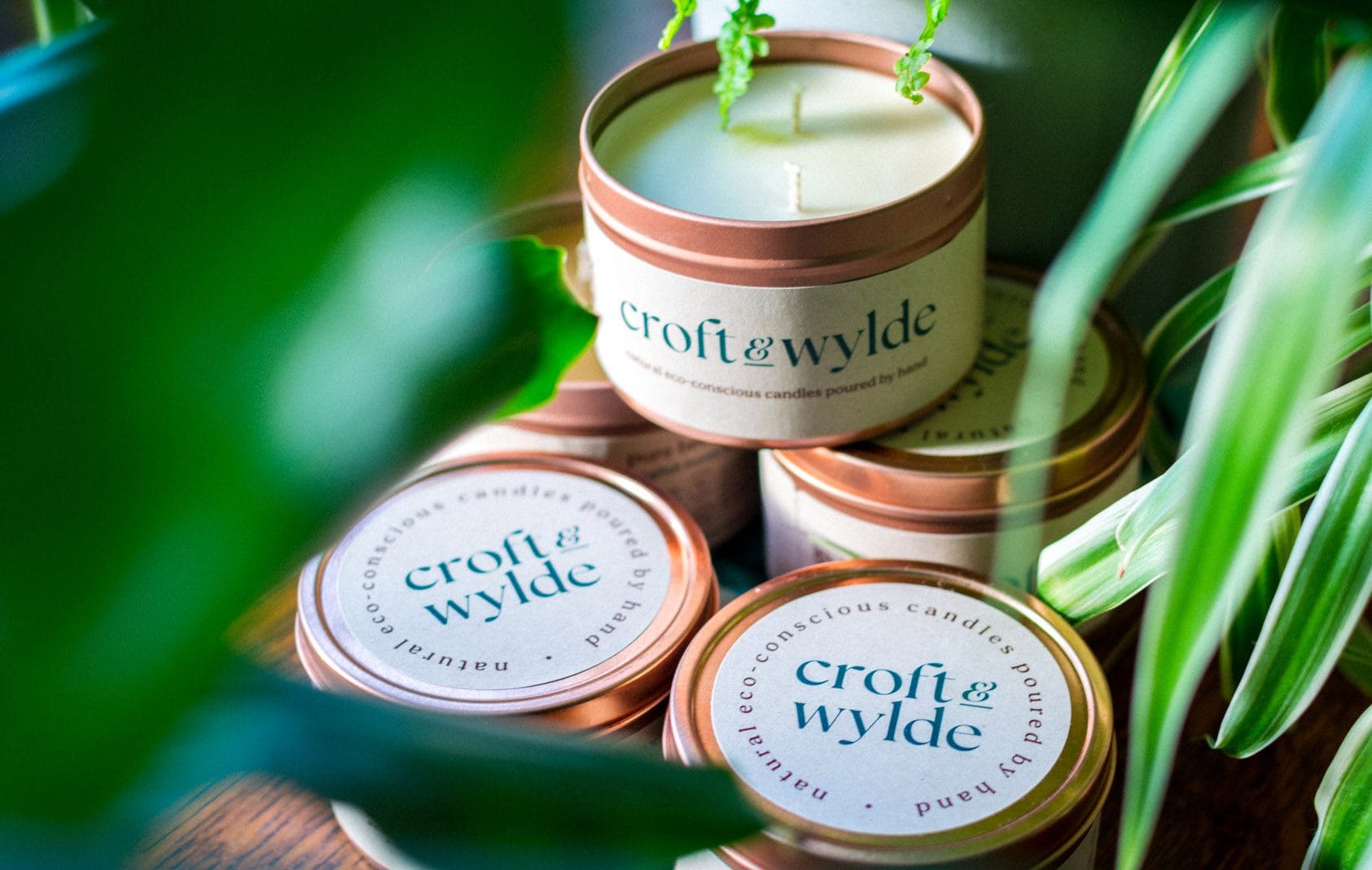 Croft & Wylde container candles in rose gold tins with kraft recycled labels that say 'Croft and Wylde - natural eco-conscious candles poured by hand' are seen pictured next to houseplant foliage on a table indoors.