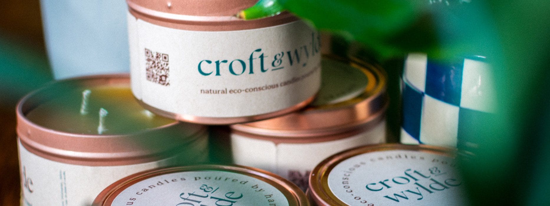 Croft & Wylde container candles in rose gold tins with kraft recycled labels that say 'Croft & Wylde - natural eco-conscious candles poured by hand' are seen pictured next to houseplant foliage on a table indoors.