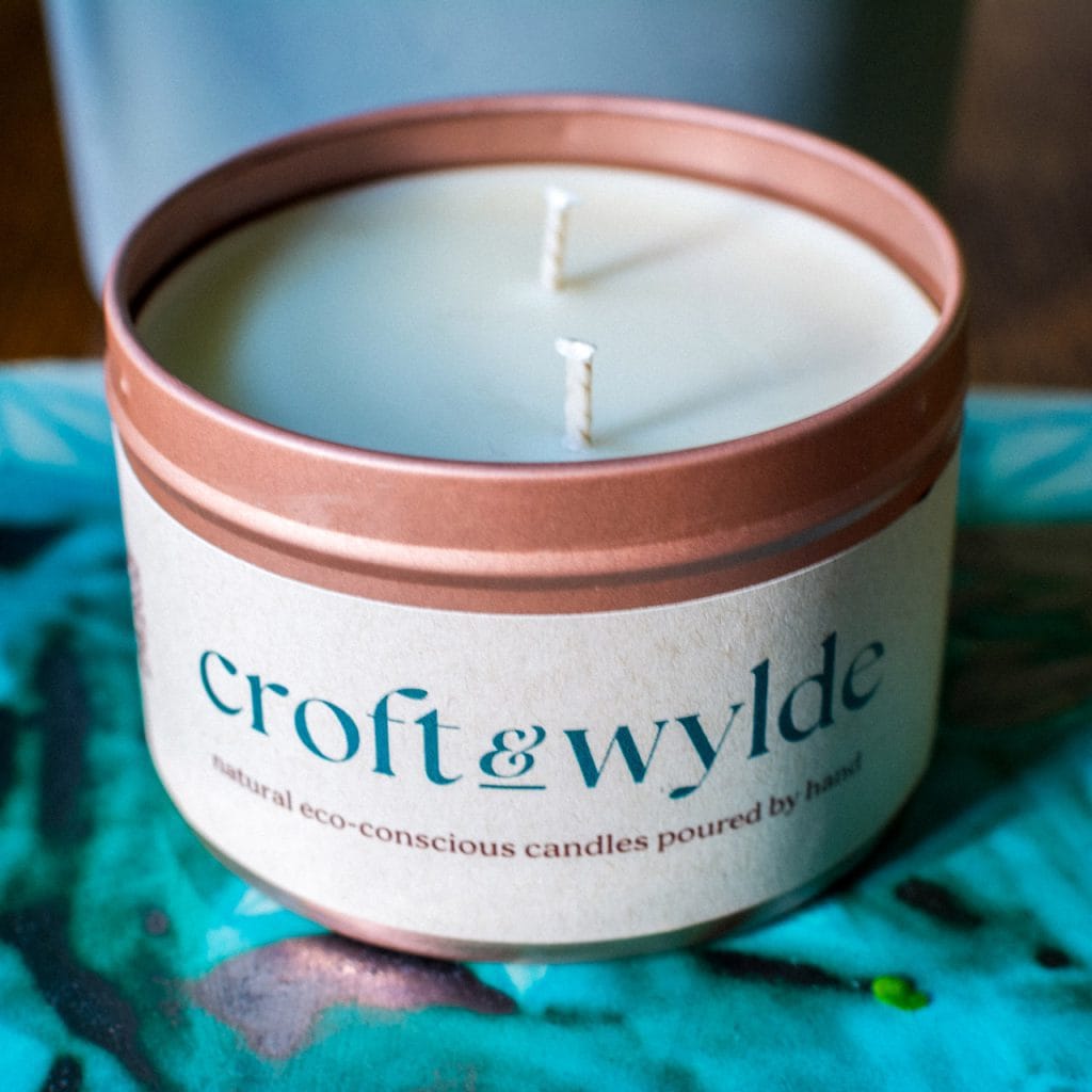 A Croft & Wylde container candle in a rose gold tin with kraft recycled labels that say 'Croft and Wylde - natural eco-conscious candles poured by hand' are seen pictured on a table indoors.