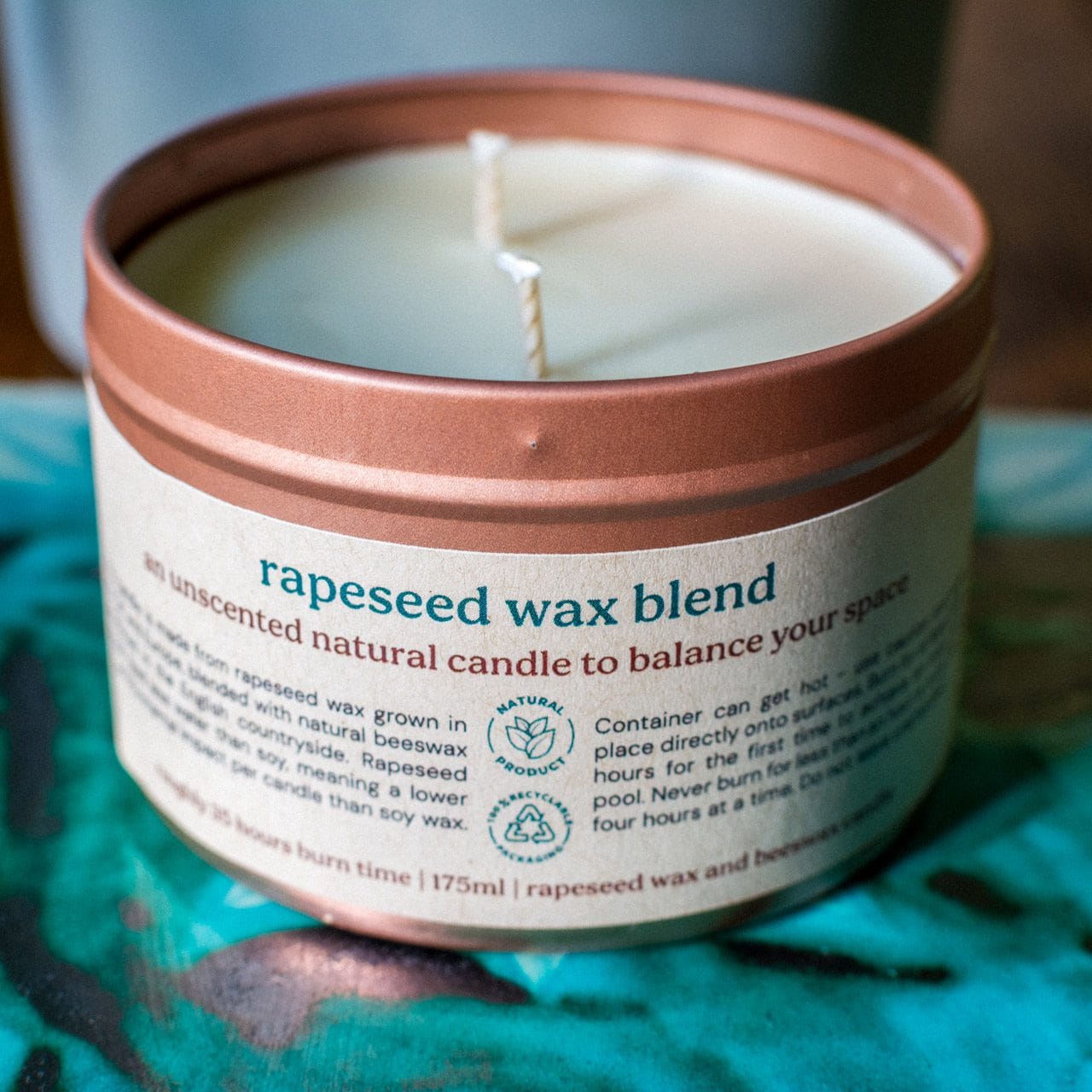 A Croft & Wylde container candle in a rose gold tin with kraft recycled labels that say 'rapeseed wax blend - an unscented natural candle to balance your space' is seen pictured on a table indoors.