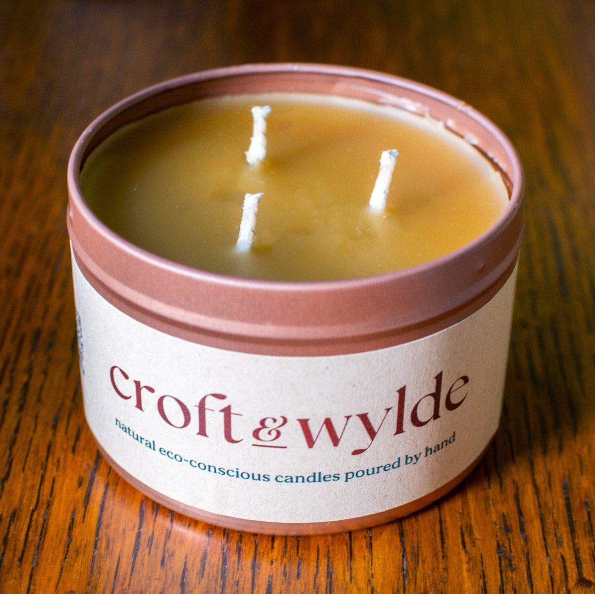 A Croft & Wylde container candle in a rose gold tin with kraft recycled labels that say 'Croft and Wylde - natural eco-conscious candles poured by hand' are seen pictured on a table indoors.