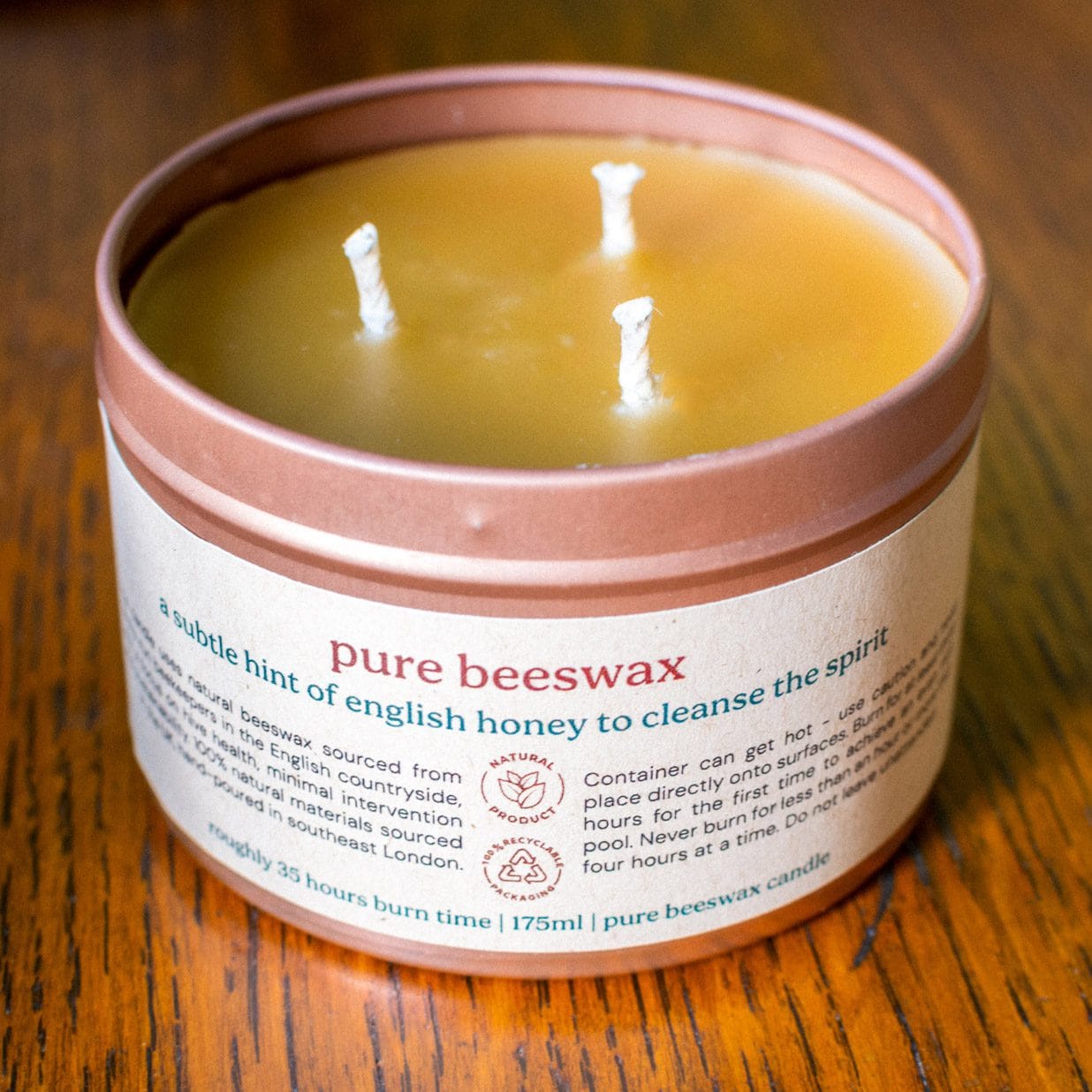 A Croft & Wylde container candle in a rose gold tin with kraft recycled labels that say 'pure beeswax - a subtle hint of english honey to cleanse the spirit' is seen pictured on a table indoors.