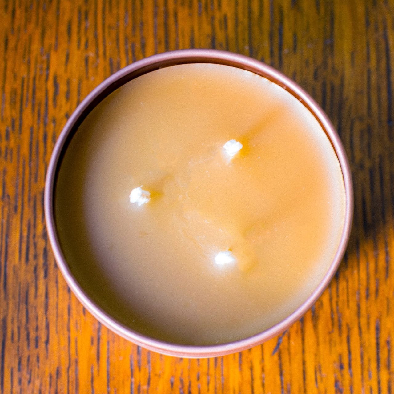 An overhead image shows the top view of a Croft & Wylde container candle in a rose gold tin with three wicks and dark caramel coloured wax.