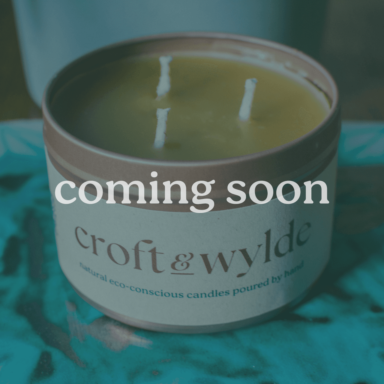 A Croft & Wylde container candle in a rose gold tin with kraft recycled labels that say 'Croft and Wylde - natural eco-conscious candles poured by hand' are seen pictured on a table indoors. The image has a blue-ish overlay with the words 'coming soon' displayed in white.