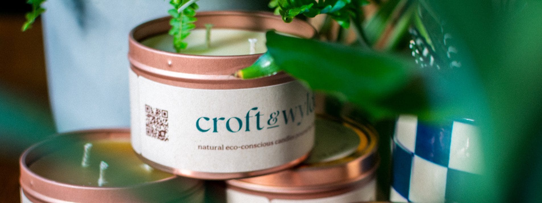 Croft & Wylde container candles in rose gold tins with kraft recycled labels that say 'Croft and Wylde - natural eco-conscious candles poured by hand' are seen pictured next to houseplant foliage on a table indoors, with a rose colour overlay casting over the left side of the image.